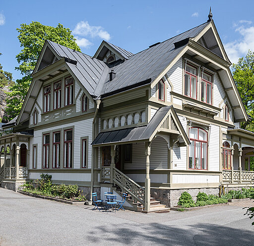 Traditionellt trähus, vit med detaljer, veranda, röda fönsterluckor, trädgårdsmöbler framför, gröna träd, soligt väder.