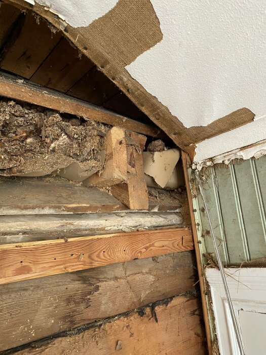 Bild på skadat tak med synliga träbjälkar, isolering, och brun väv som hänger. Slitage eller vattenskada syns.