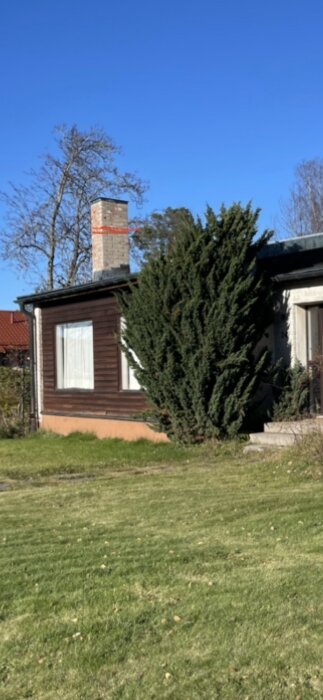 Ett brunt hus med vit dörr, skorsten, en stor buske, gräs och klarblå himmel.