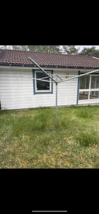 Rotary clothesline framför en vit trähusvägg, fönster, veranda, övervuxen gräsmatta, träd i bakgrunden.