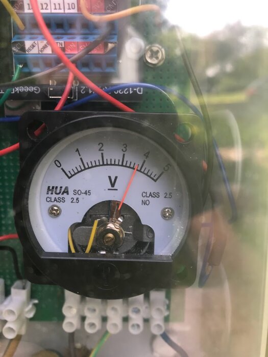 Voltmeter i en elektrisk krets, anslutna kablar, genomskinlig plastkåpa, grön kretskortsplatta i bakgrunden.