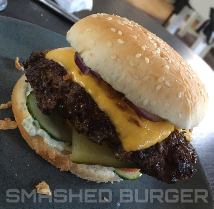 En cheeseburger med lök, cheddarost, pickles, sås, sesamfröbröd på en tallrik, text "SMASHED BURGER" nedanför.