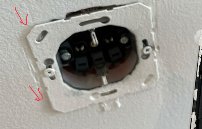 En avlägsnad eluttag's täckplatta, exponerad kabelanslutning, vita väggen, röda pilar indikerar problemområden.