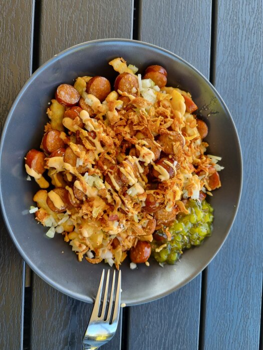 En tallrik med mat: korv, potatis, lök, sås och krispiga lökbitar, med en gaffel, på ett mörkt träbord.