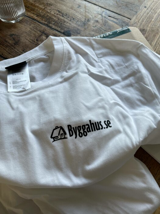 Vit t-shirt med logotyp "Byggahus.se" och en liten hus-ikon, ligger på träyta.