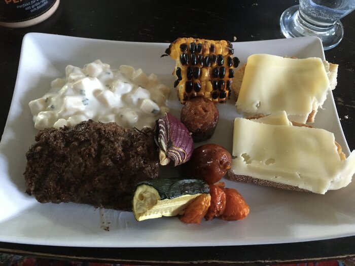 En tallrik med blandad mat: köttfärslimpa, majskolv, potatissallad, grillade grönsaker och smörgås med ost.
