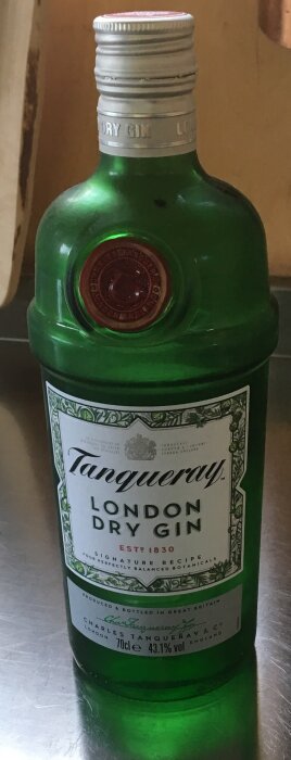 Grön flaska med etikett, Tanqueray London Dry Gin, stående på metallisk yta.