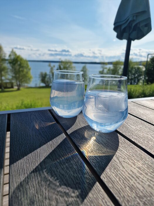 Två vattenglas på träbord med sjöutsikt, soligt, grönska, parasollskugga.