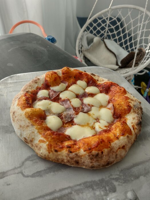 Hembakad pizza med tomatsås, ost, korv på bakplåtspapper, otydlig bakgrund inne.