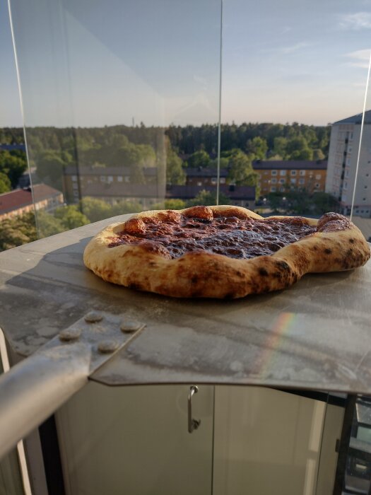 Pizza på en balkong med reflekterande glasräcke och bakgrund av byggnader och träd i solljus.