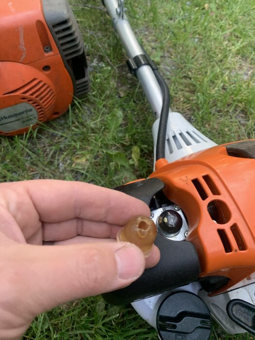 Hand håller genomskinlig plastbricka framför röd trimmer på gräsmatta. Reparation eller underhåll kan antydas.