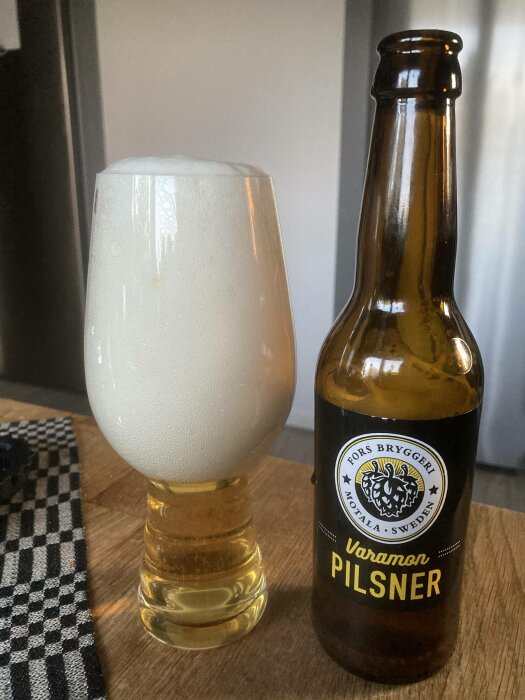 Ölglas med överflödigt skum bredvid halvtom pilsnerflaska på träbord.