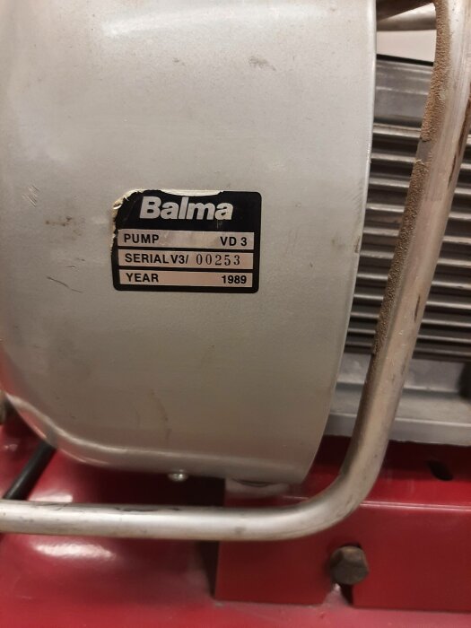 Sliten märkplåt på maskin, "Balma", pumpmodell, serienummer, tillverkningsår 1989. Röd del av maskin synlig.