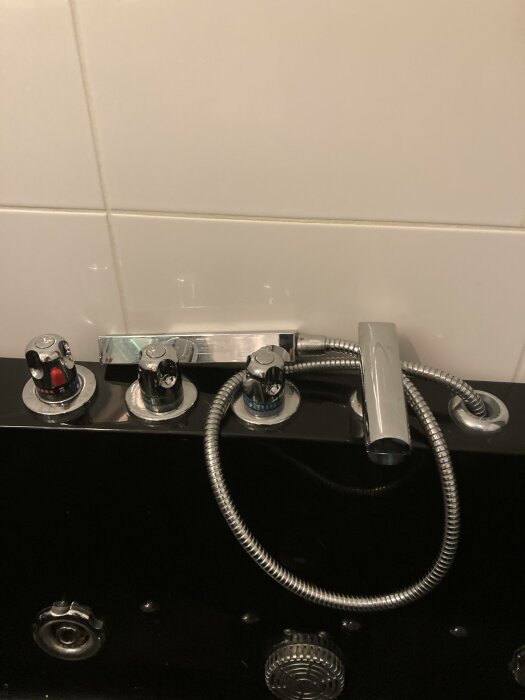 Badkarsblandare med röda och blåa markeringar, handdusch, svart badkar, vita kakelväggar.