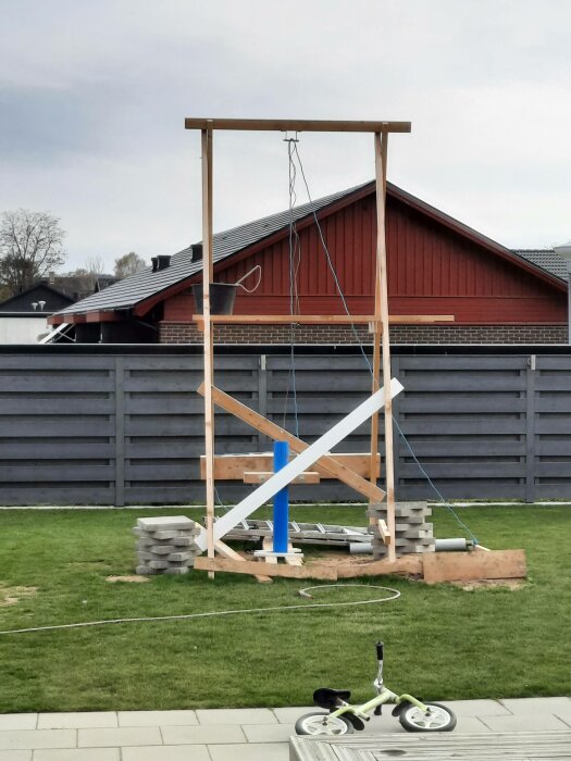 Träkonstruktion, rep, hink, betongplattor, barnsparkcykel, gräs, grå staket, röd byggnad i bakgrunden.