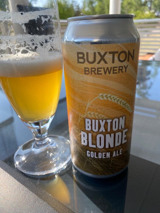 Burk med Buxton Blonde Golden Ale vid sidan av ett halvfullt ölglas, soligt utomhus.