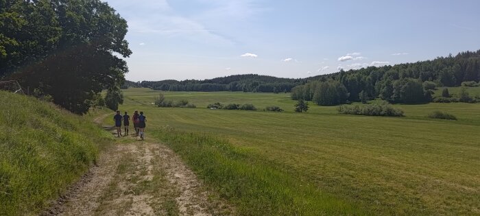 Personer vandrar på stig genom soligt, lummigt landskap med gröna fält och skogar.