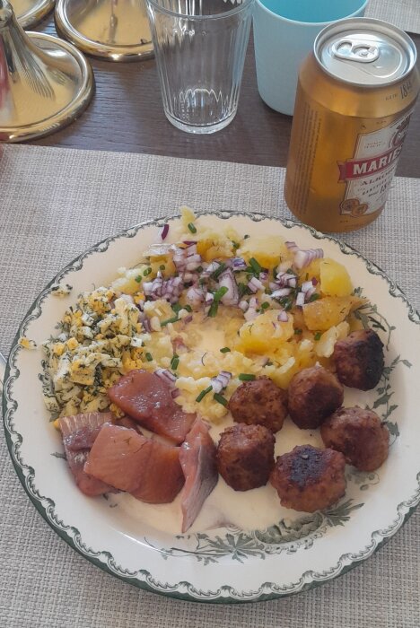 En tallrik med potatis, köttbullar, rökt skinka, äggröra, gräddsås, hakad rödlök, dekorerad tallrik, ölburk bredvid.