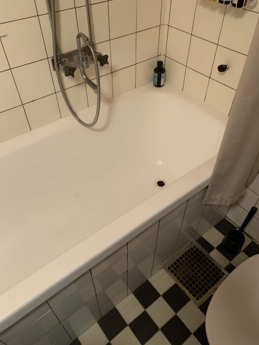 Ett tomt badkar med duschmunstycke, svartvita kakelplattor och en flaska vid hörnet.