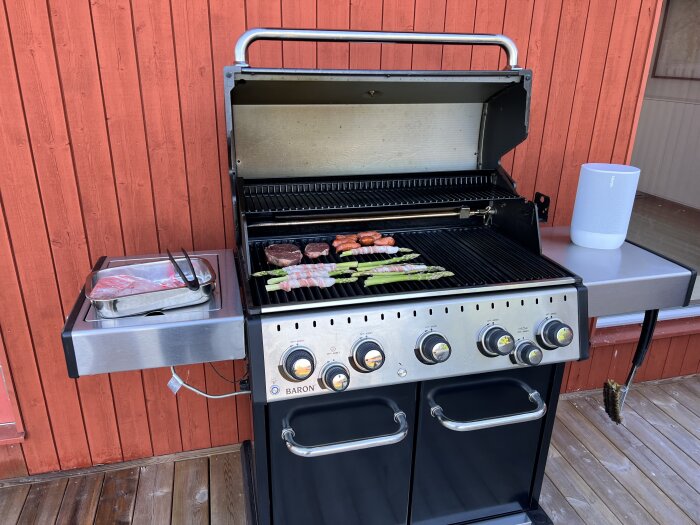 Gasolgrill med kött, sparris på grillen, röd trävägg i bakgrunden, soligt ute.