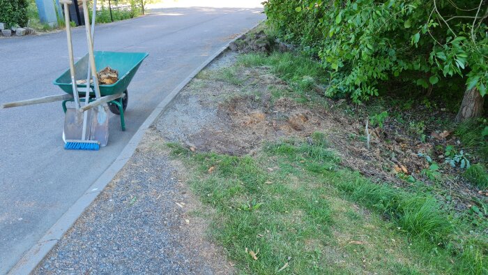 Grön skottkärra med trädgårdsverktyg, skadad gräsmatta, kantsten, asfalterad väg, gröna buskar, soligt utomhus.