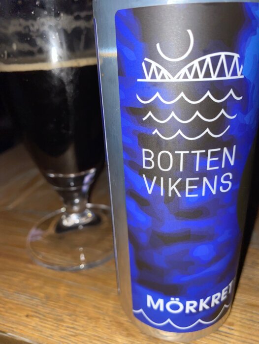 Ölburk med text "BOTTENVIKENS MÖRKRET", grafisk design av bro, vågor och måne, delvis synligt ölglas bakom.