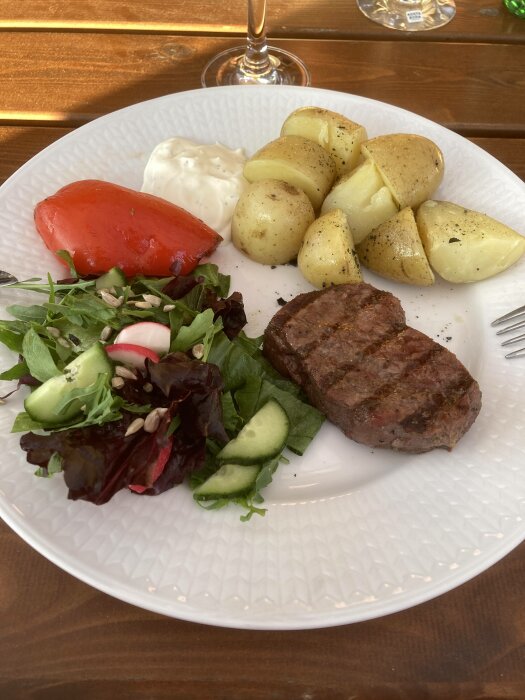 En tallrik med grillad köttbit, kokt potatis, sallad, tomat och vit sås.