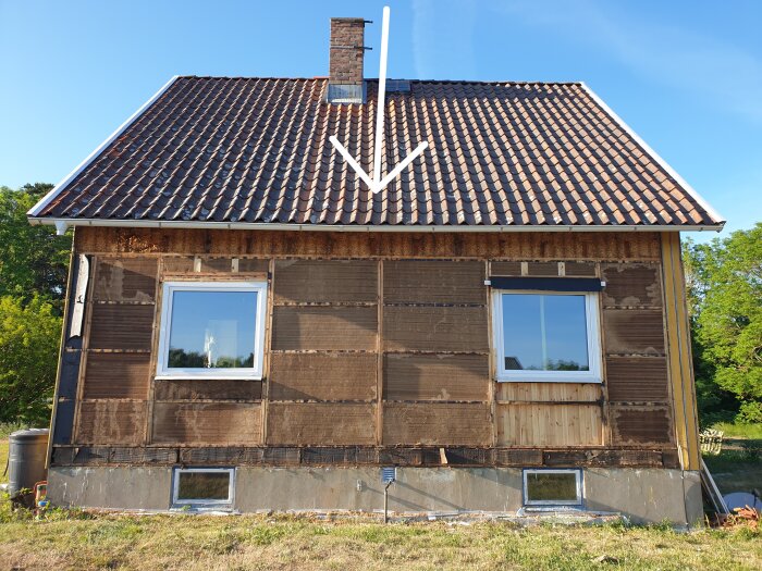 Tvåvånings trähus med tegeltak, skorsten, saknar yttre väggpaneler, renoveringsarbete synbart, grön omgivning, klar himmel.