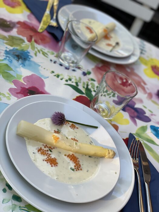 En tallrik med sparris och sås, blomsterduk, tomma glas, kvarlämnade tallrikar, utomhusmåltid.