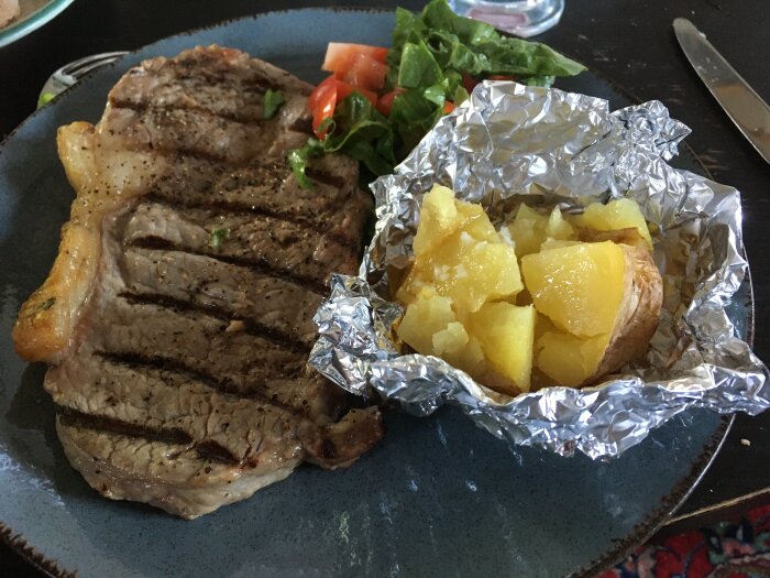 Grillad biff med grillränder, bakad potatis i folie, sallad på tallrik, matservering inomhus.