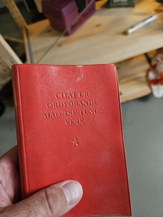 Röd bok i hand, "Citat ur ordförande Mao Tse-tungs verk" på omslaget, verktyg och hyllor i bakgrunden.