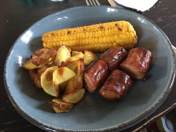 Grillad majskolv, rostade potatisklyftor och glaserade, grillade köttbitar på blå tallrik.