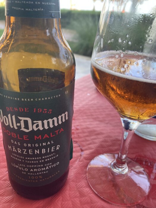 Ölflaska och halvfyllt glas på röd duk utomhus. Märke: Doble Malta Märzenbier. Etikett på spanska. Avslappnad atmosfär.