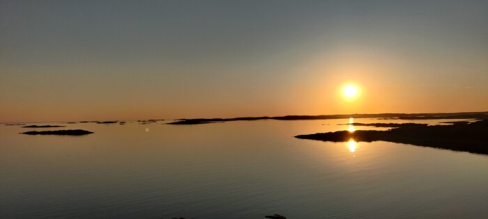 Solnedgång över lugnt hav, små öar, avspeglad sol, vidsträckt utsikt, orörd natur, gyllene himmel, stämningsfullt.