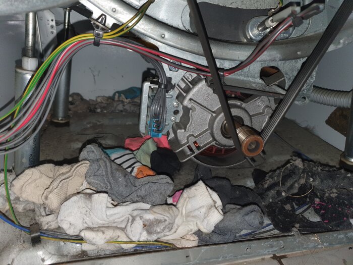 Tvättmaskinens insida med kläder och smuts runt trumman och motorn. Kablar och mekaniska komponenter synliga.