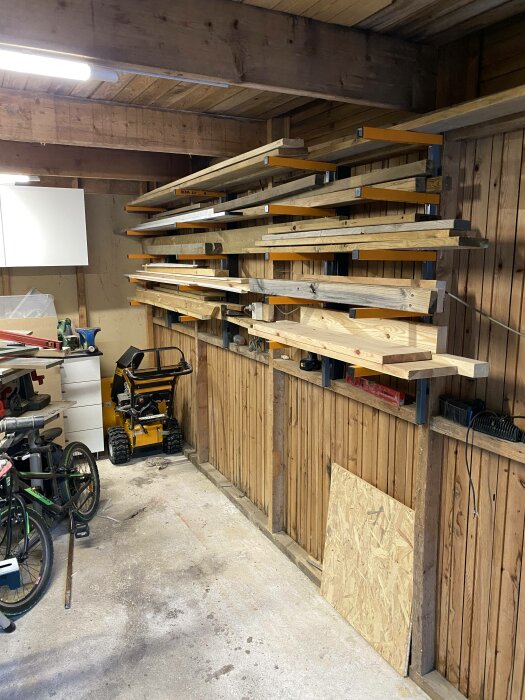 Garage med virkeshyllor, gräsklippare, cykel, verktyg och spånplatta mot träväggar och betonggolv.