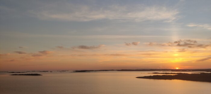 Solnedgång över havet, himlen i orange toner, stilla vatten, små öar och holmar, fredfullt och vackert.