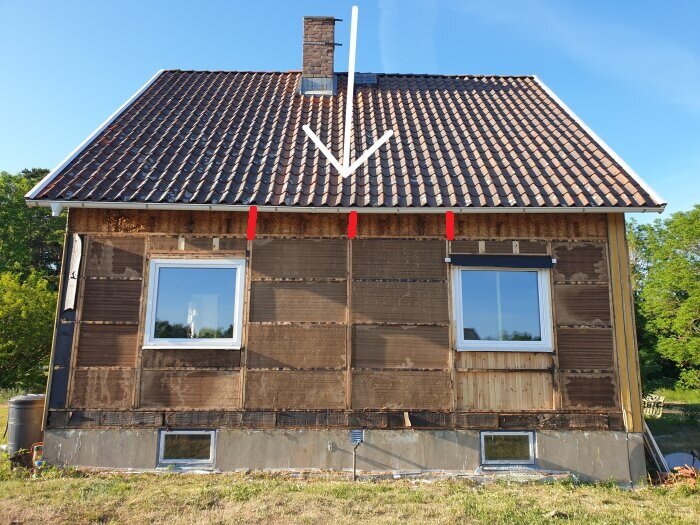 Hus under renovering, fasaden saknar panel, exponerar isoleringsmaterial, grönt gräs, blå himmel, två fönster.