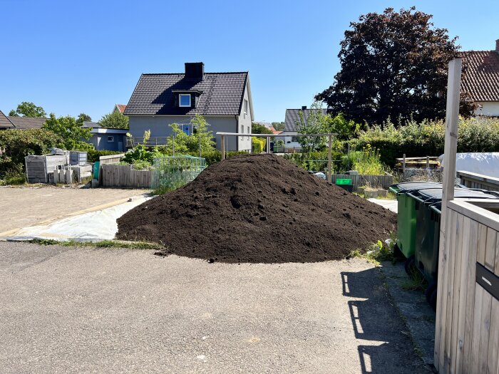 Stor hög med mörk jord, bostadshus i bakgrunden, solig dag, gröna avfallskärl, asfalterad yta framför.