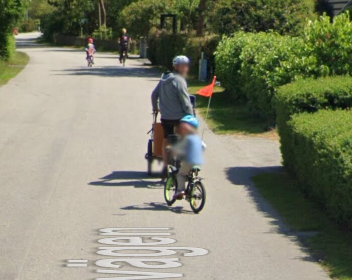 Person cyklar med barn i cykelkärra, andra cyklister i bakgrunden, solig dag.