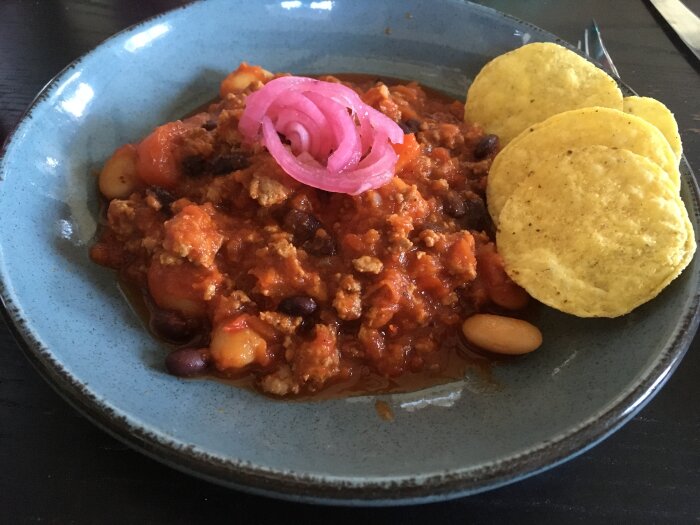 Tallrik med chili con carne, rödlök, nachochips, på mörkt bord.