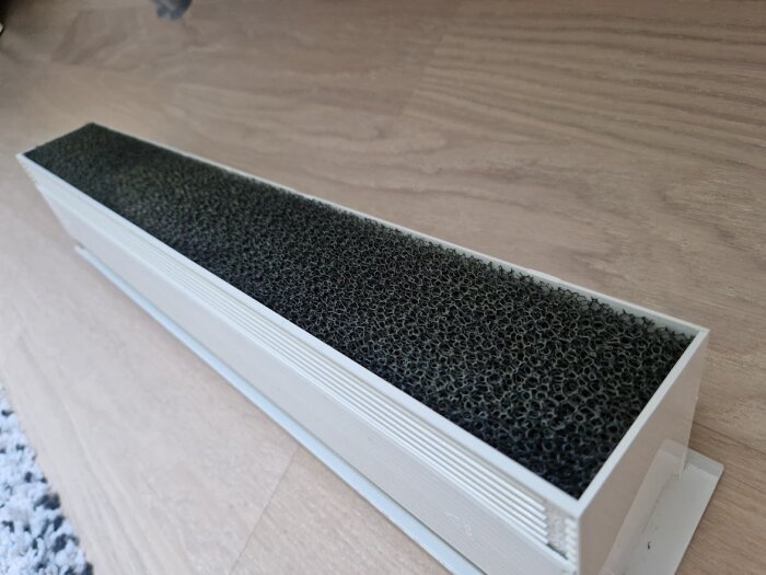 HEPA-filter i vit plastbehållare på ett trägolv, används i luftrenare eller ventilationssystem.