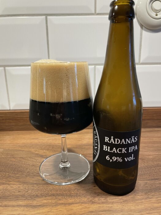 En mörk IPA-öl med tjockt skum i glas bredvid öppnad flaska.