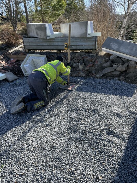 Arbetare i säkerhetskläder knäböjer på grus, jobbar vid byggarbetsplats med sten och betongelement.