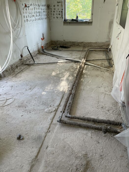Rörledningar exponerade i tomt rum under renovering, betonggolv, kabelhärvor, soligt fönster.
