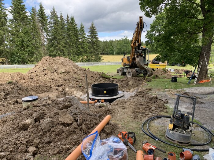 Grävmaskin jobbar, högar med jord, avloppsledningar, verktyg på byggarbetsplats, gröna träd i bakgrunden.