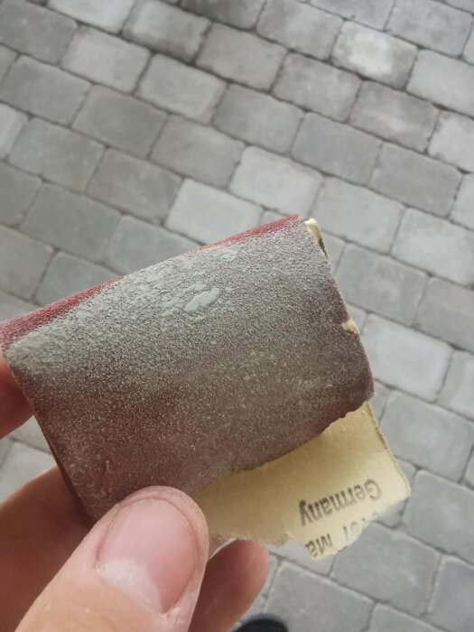 Hand håller slitet sandpapper, rödbrun yta, bakgrund av trottoarstenar, närbild, utomhus, dagsljus.