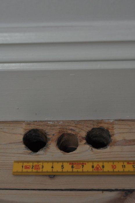 Tre huller i et fyrretræsgulv, hver med en diameter på 20 mm, med en lineal som måleredskab nær ved.