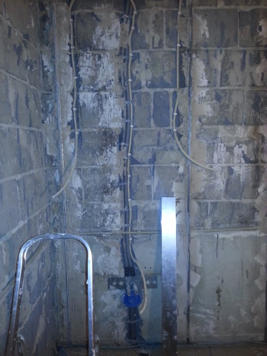 Badrumsvägg med blåbetong, visa monterade rör och ojämn yta, stege till vänster och metallverktyg som mäter avvikelser i väggen.