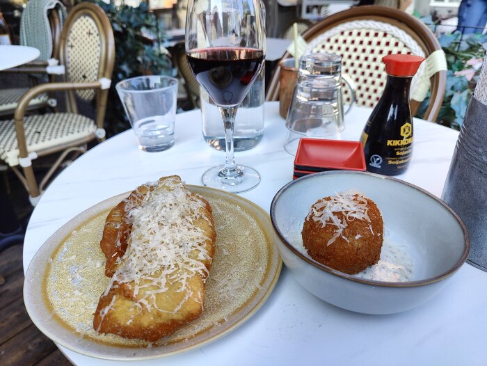 Mat på utomhusbord: arancini, toast med ost, sojasås, rödvin, vattenglas. Avslappnat caféstämning.